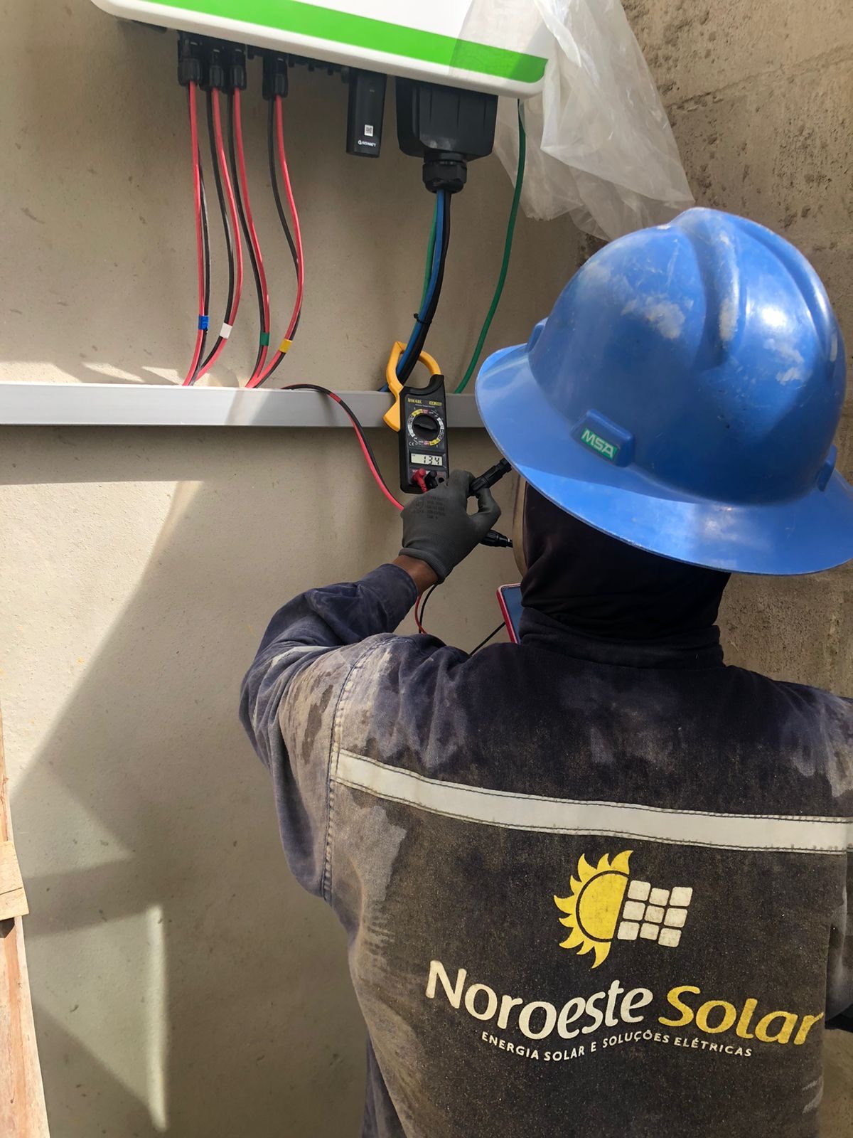 Técnico da Noroeste Solar com capacete e luvas medindo o inversor com alicate amperímetro